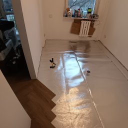 ANDRZEJ DAMPC SZYBKA USTERKA I REMONT - Fragment układania parkietu w jodełkę, widoczna folia izolacyjna, młotek i fragment okna z przedmiotami. Ujęcie z góry, z perspektywy osoby stojącej w pomieszczeniu.