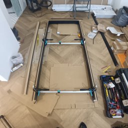 ANDRZEJ DAMPC SZYBKA USTERKA I REMONT - Rama drzwiowa w trakcie montażu na podłodze w jodełkę, zabezpieczona kartonem. Widoczne narzędzia, skrzynka narzędziowa i drabina w tle. Ujęcie z góry.