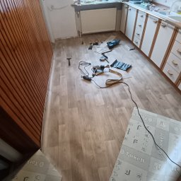 ANDRZEJ DAMPC SZYBKA USTERKA I REMONT - Kuchnia w trakcie układania paneli podłogowych. Widoczne narzędzia, częściowo ułożone panele Tarkett i fragment boazerii na ścianie. Ujęcie z góry, widać stopy osoby robiącej zdjęcie.
