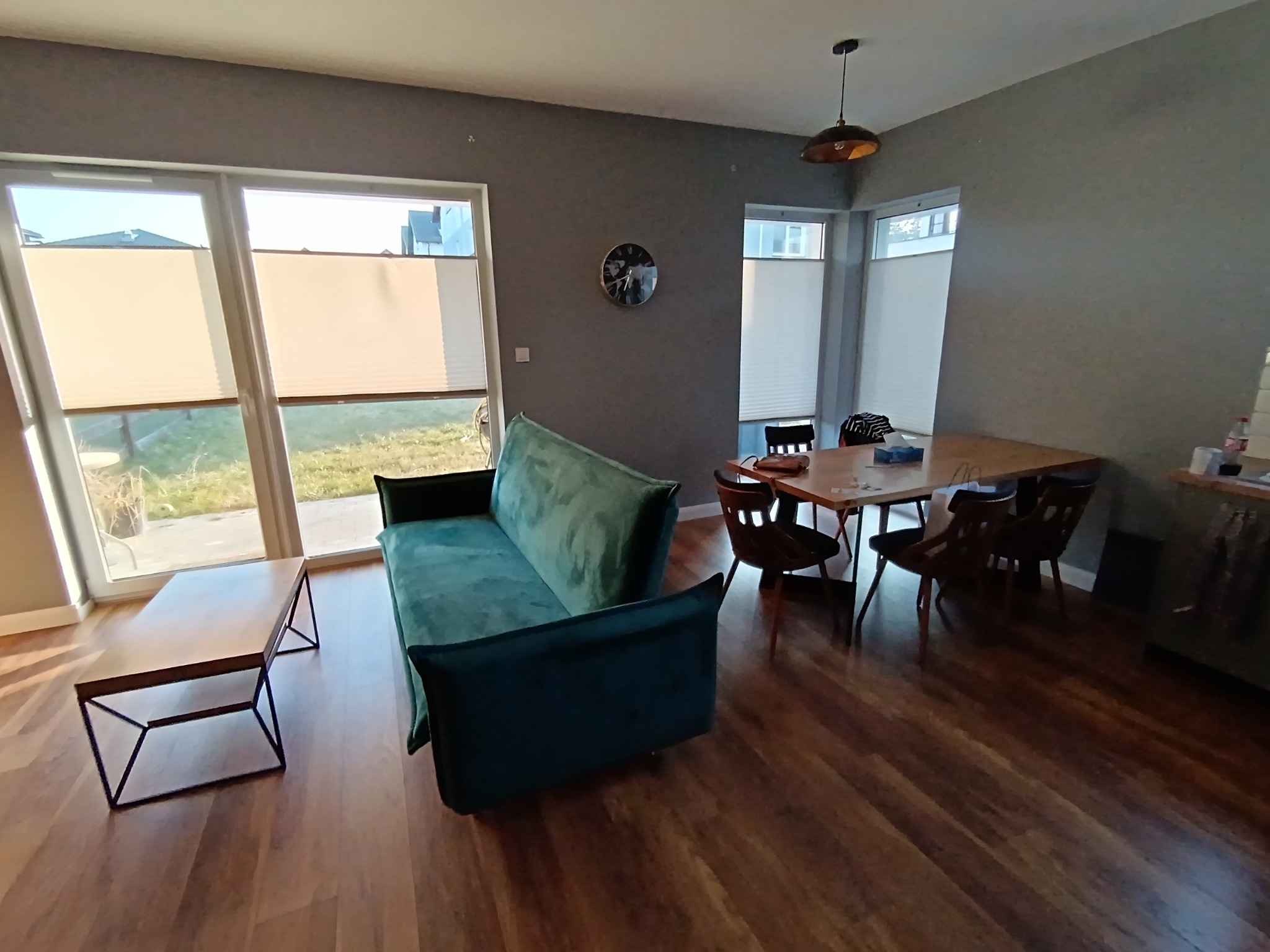 Nowoczesne wnętrze: parkiet, szary salon z zieloną sofą, stolikiem kawowym i stołem jadalnym. Okna z roletami plisowanymi wpuszczają jasne światło.