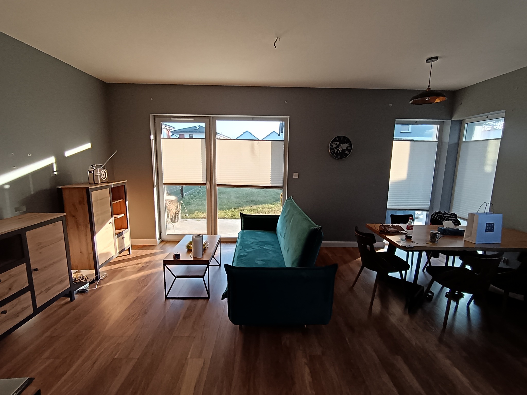 Jasne wnętrze pokoju dziennego po remoncie: szare ściany, drewniana podłoga, nowoczesne meble, okna z żaluzjami i lampa wisząca. Widok na odnowione pomieszczenie.