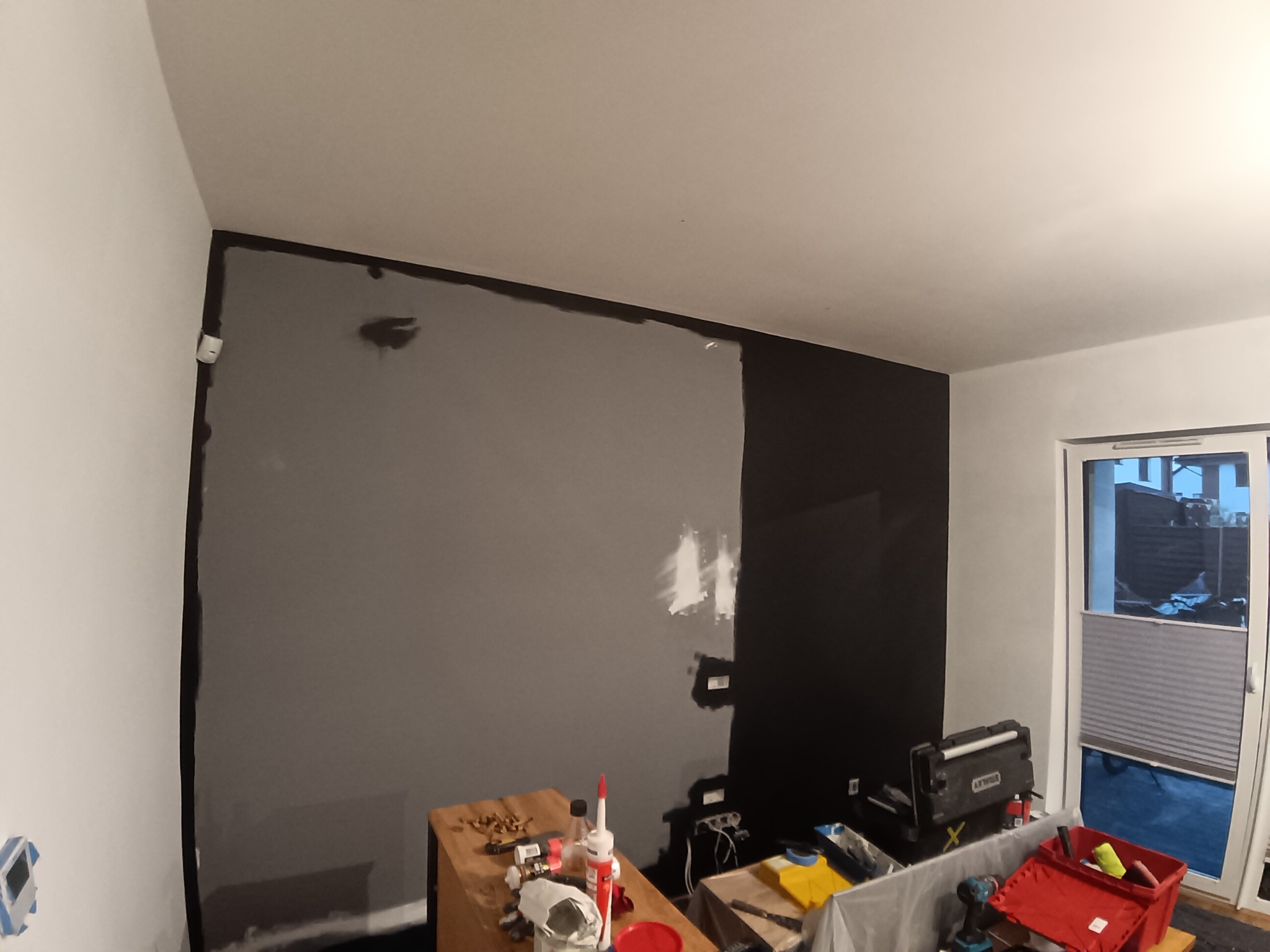 Ściana w trakcie malowania na szaro i czarno, widoczne narzędzia malarskie, częściowo pomalowana ściana, listwy przypodłogowe, okno balkonowe.