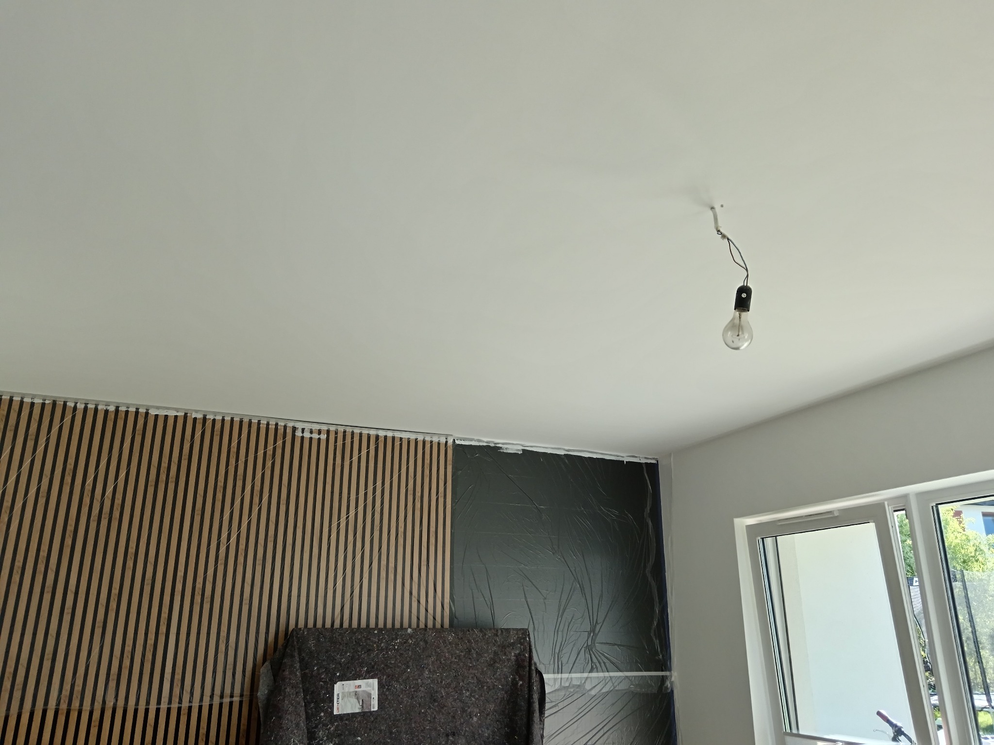 Montaż paneli ściennych: widok na fragment ściany z zamontowanymi panelami drewnianymi, zabezpieczonymi folią, oraz sufit z wiszącą żarówką. Widoczne okno.