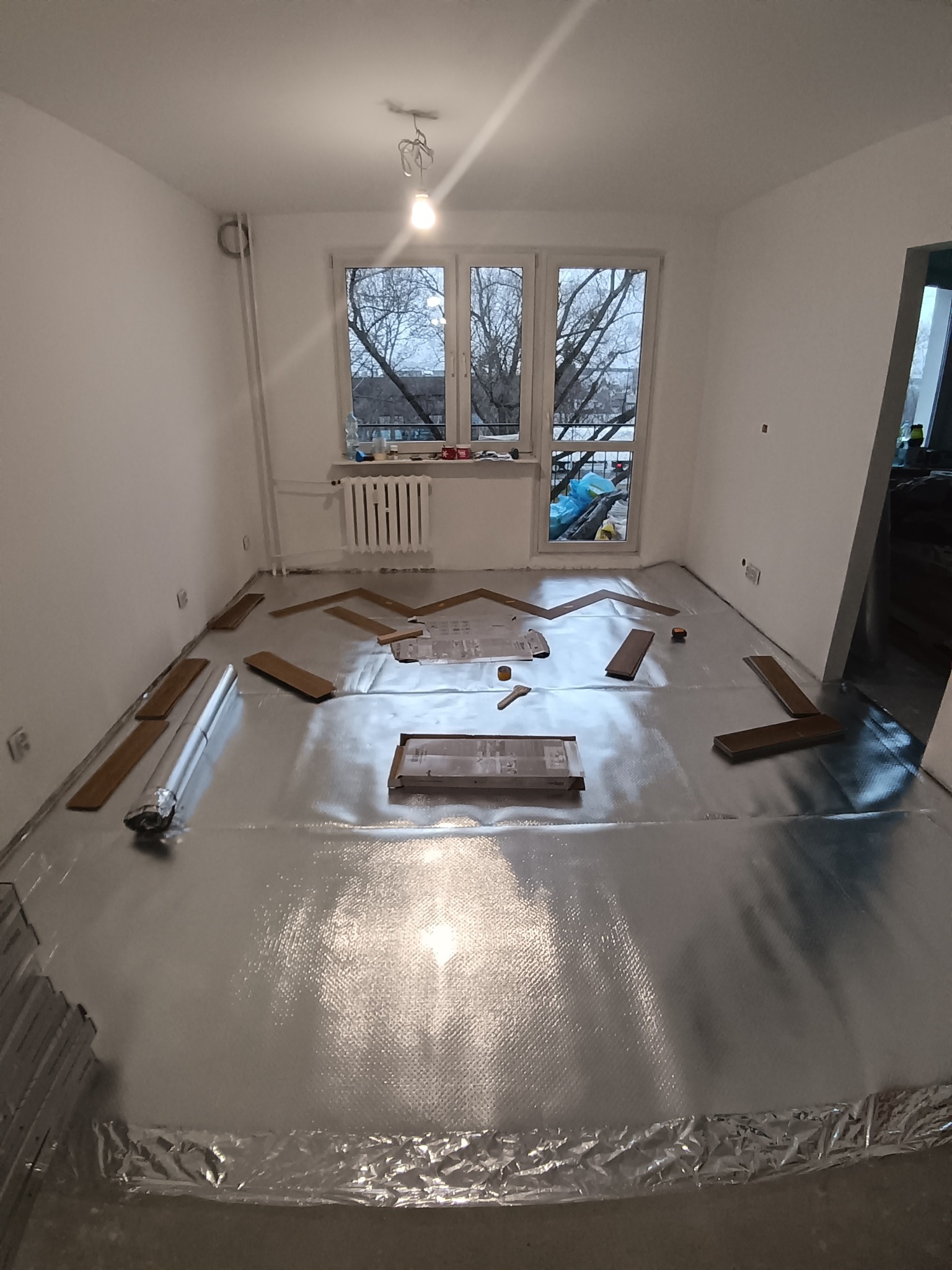 Przygotowanie do montażu paneli podłogowych: rozłożona folia izolacyjna, panele, narzędzia w jasnym pokoju z oknem. Widok z góry, etap prac przed ułożeniem całości.