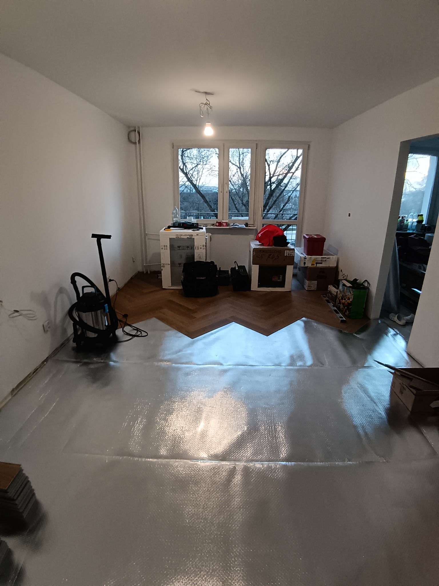 Rozłożona folia izolacyjna i fragment ułożonych paneli w jodełkę w jasnym pokoju. Widoczny odkurzacz przemysłowy i okno z widokiem na drzewa.