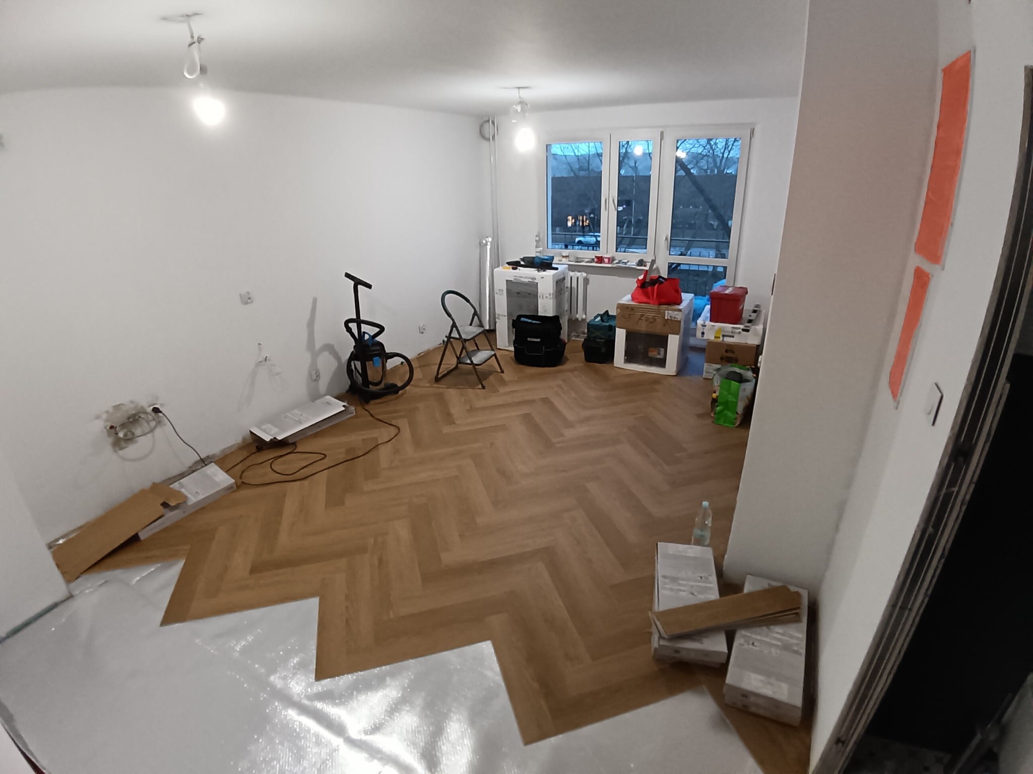 Fragment układania parkietu w jodełkę w jasnym pokoju. Część podkładu widoczna, reszta podłogi wykończona. W tle okno i sprzęt budowlany.