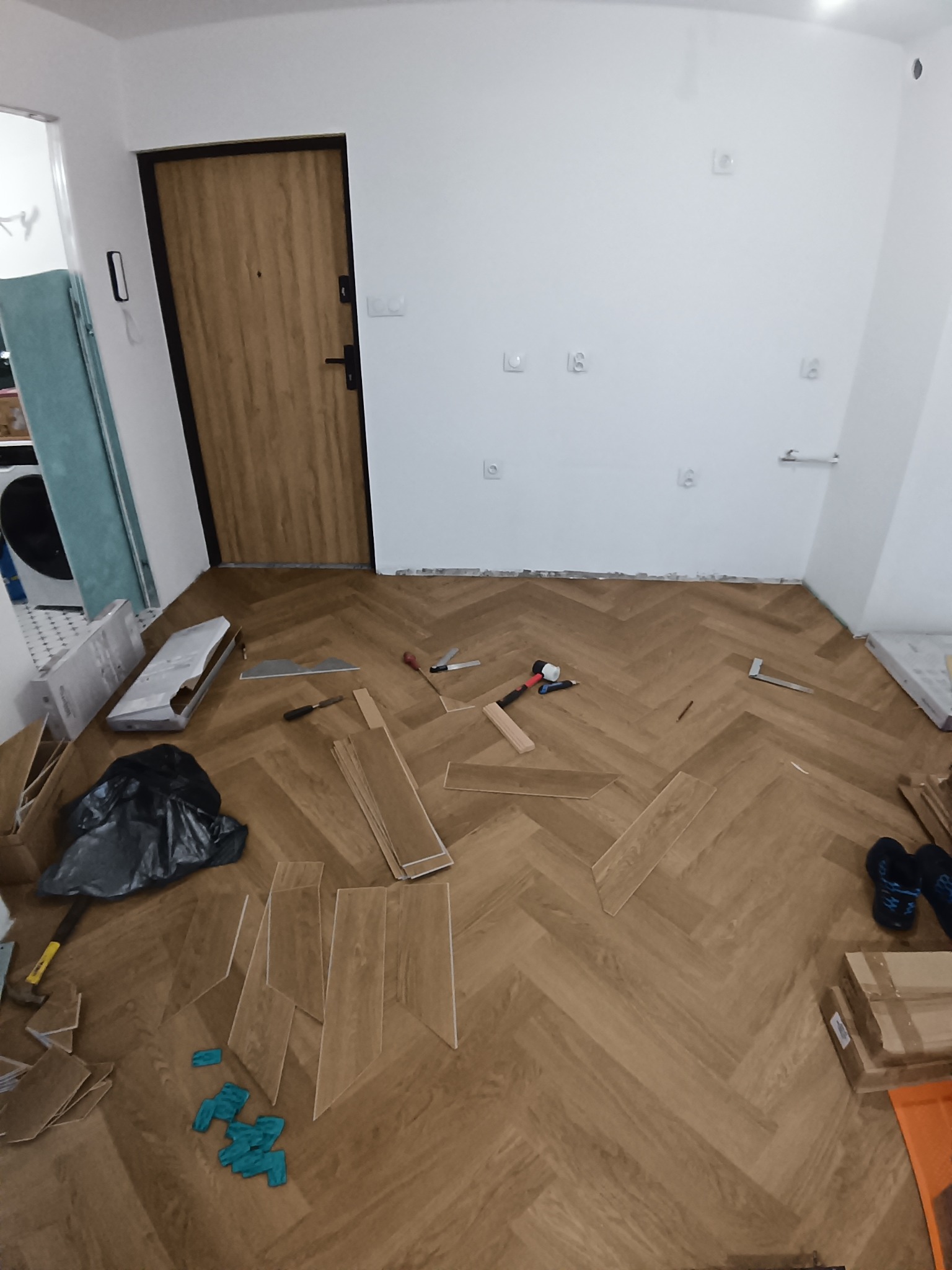 Układanie paneli w jodełkę: fragment podłogi z ułożonymi panelami, narzędzia pracy, paczki paneli i listwy przypodłogowe przy ścianie. Widok z góry.