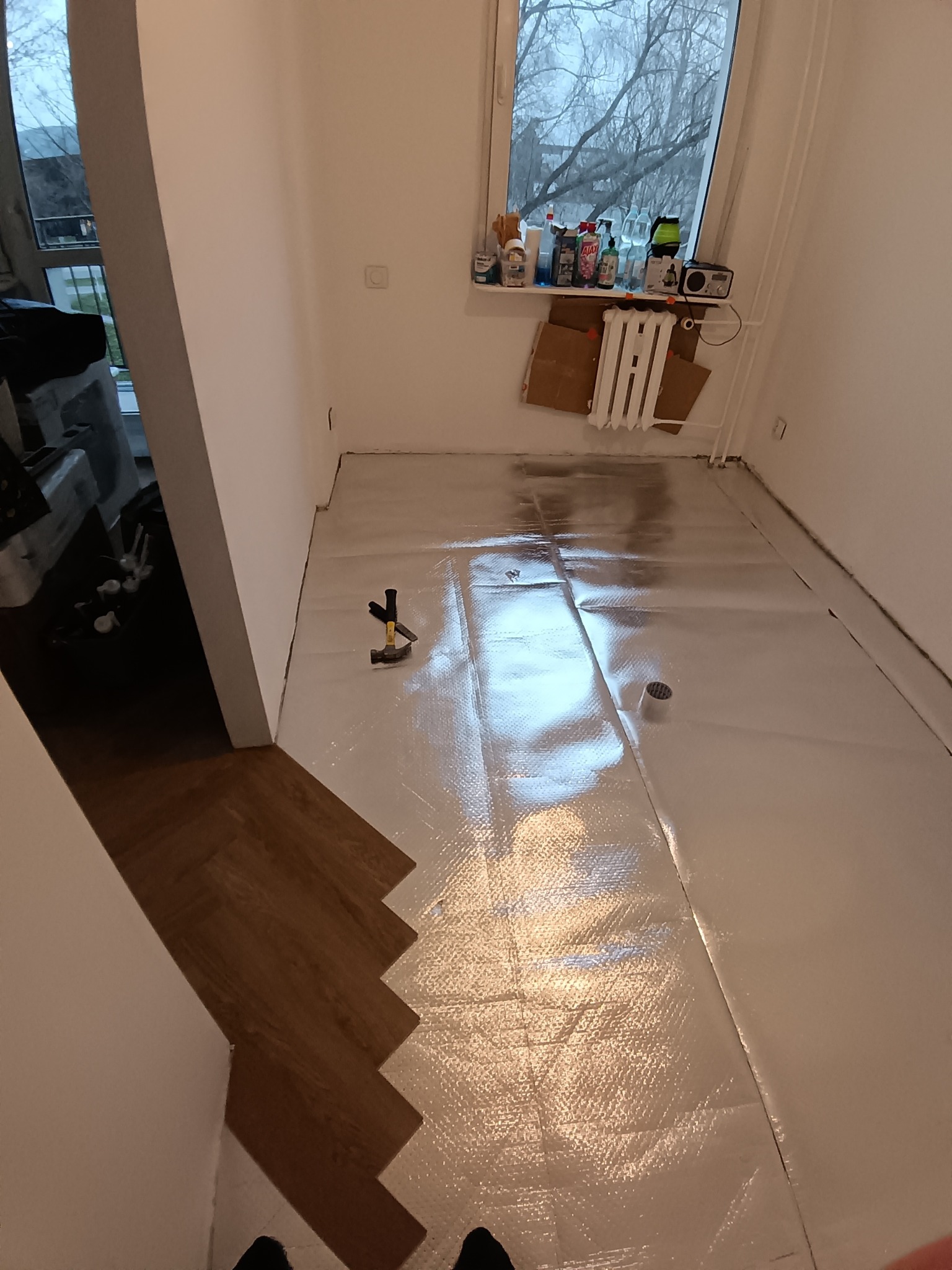 Fragment układania parkietu w jodełkę, widoczna folia izolacyjna, młotek i fragment okna z przedmiotami. Ujęcie z góry, z perspektywy osoby stojącej w pomieszczeniu.