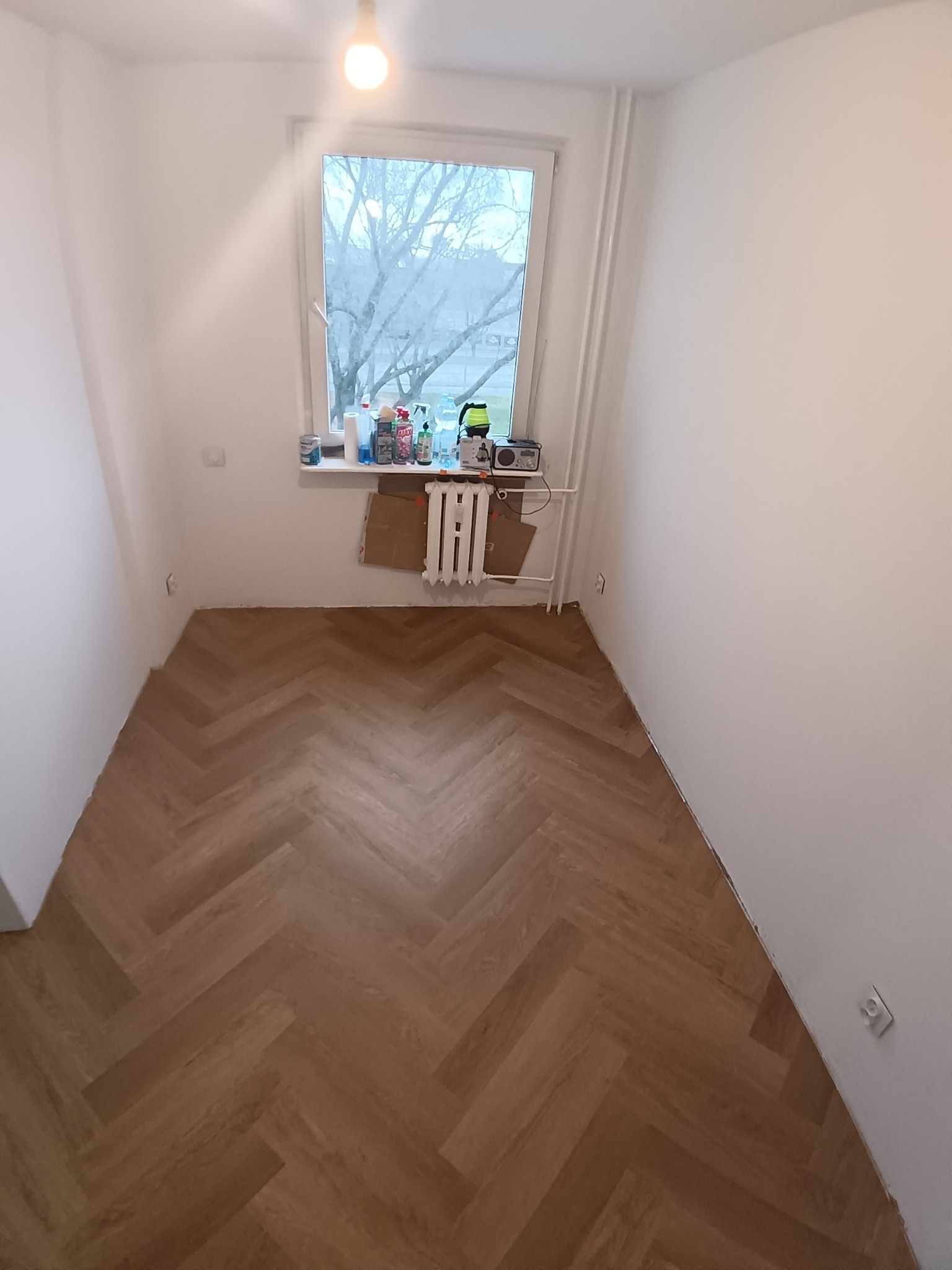 Jasne panele ułożone w jodełkę w pokoju z białymi ścianami i oknem. Na parapecie okna widoczne różne przedmioty. Grzejnik pod oknem częściowo zasłonięty kartonem.