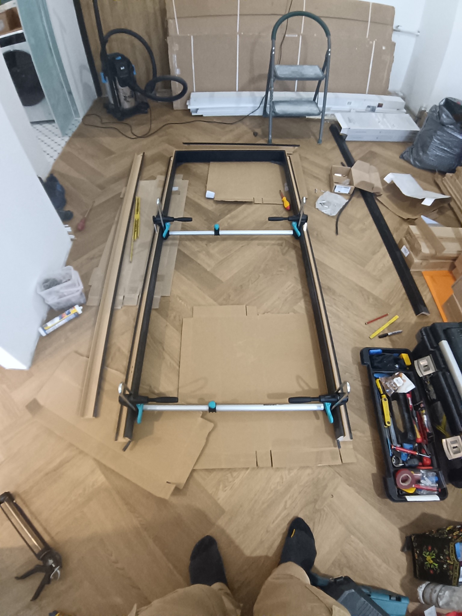Rama drzwiowa w trakcie montażu na podłodze w jodełkę, zabezpieczona kartonem. Widoczne narzędzia, skrzynka narzędziowa i drabina w tle. Ujęcie z góry.