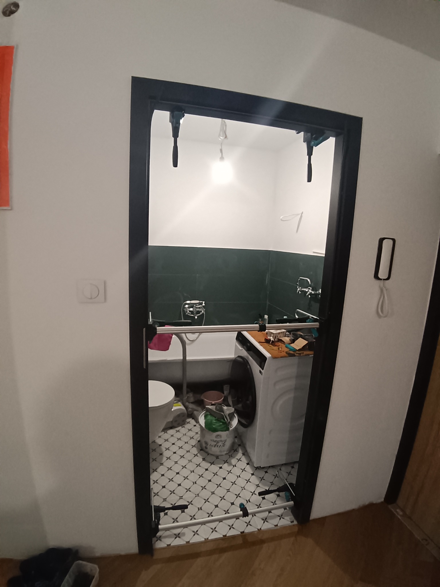 Ciemna rama drzwi do łazienki z widoczną wanną, pralką i toaletą. W górnej części ramy zamontowane narzędzia. Wnętrze w trakcie prac, widoczne akcesoria.