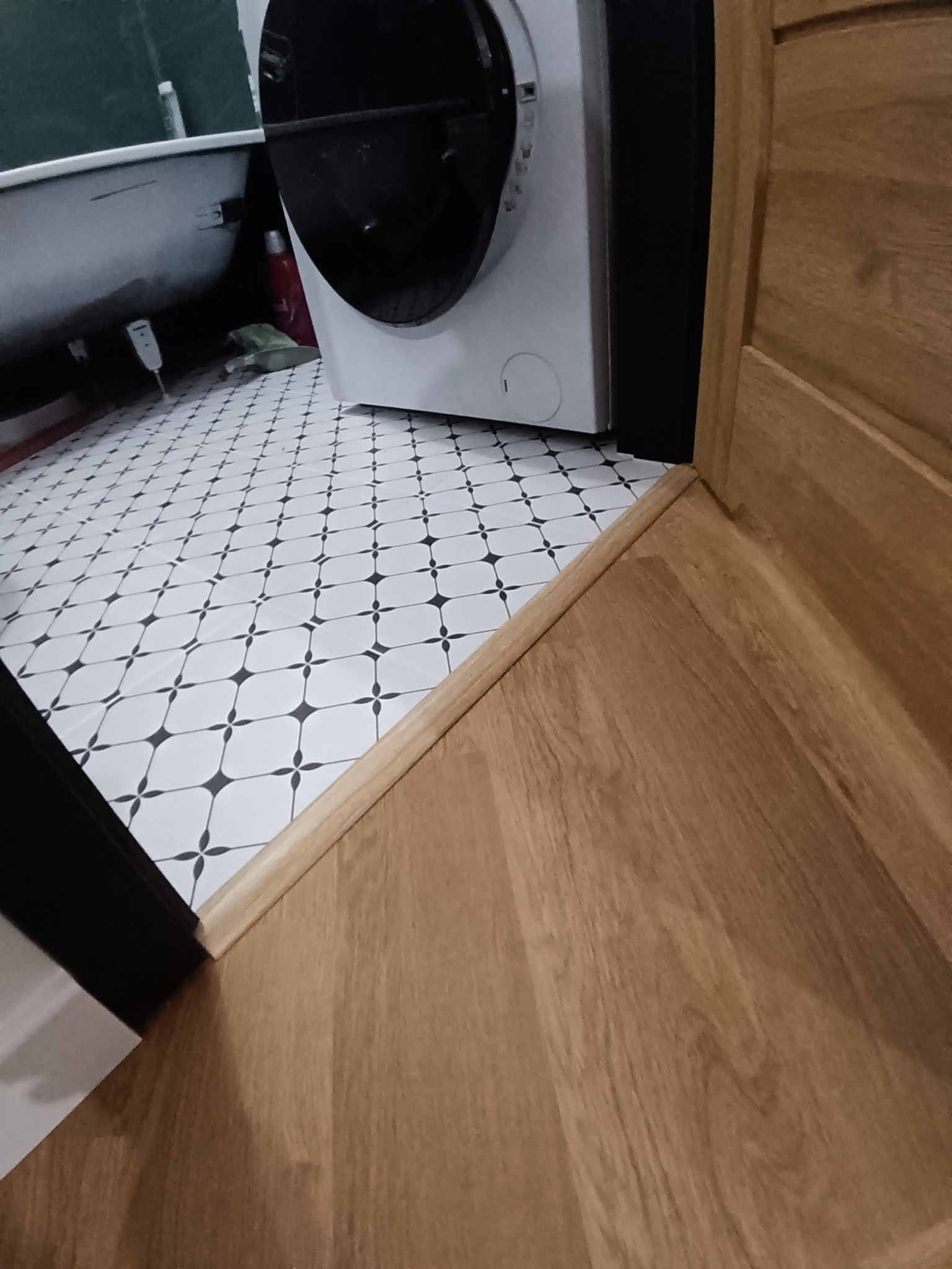 Biało-czarna podłoga w łazience z wzorem, sąsiadująca z drewnianą podłogą i fragmentem pralki. Widoczny próg łączący dwa rodzaje podłóg.