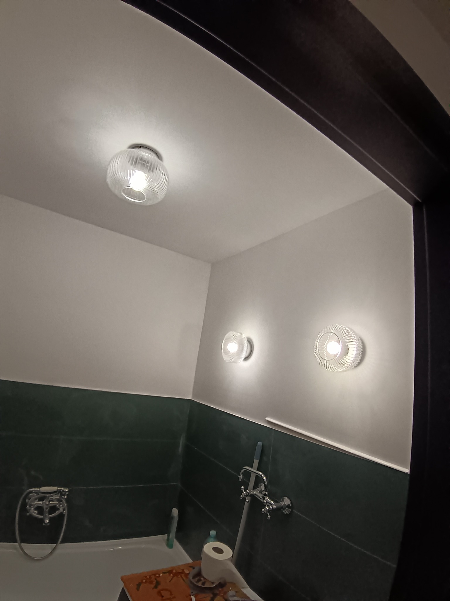 Nowoczesna łazienka z zielonymi płytkami, wanną i trzema okrągłymi lampami sufitowymi. Widok zza framugi drzwi, oświetlenie ciepłe, tworzące przytulną atmosferę.