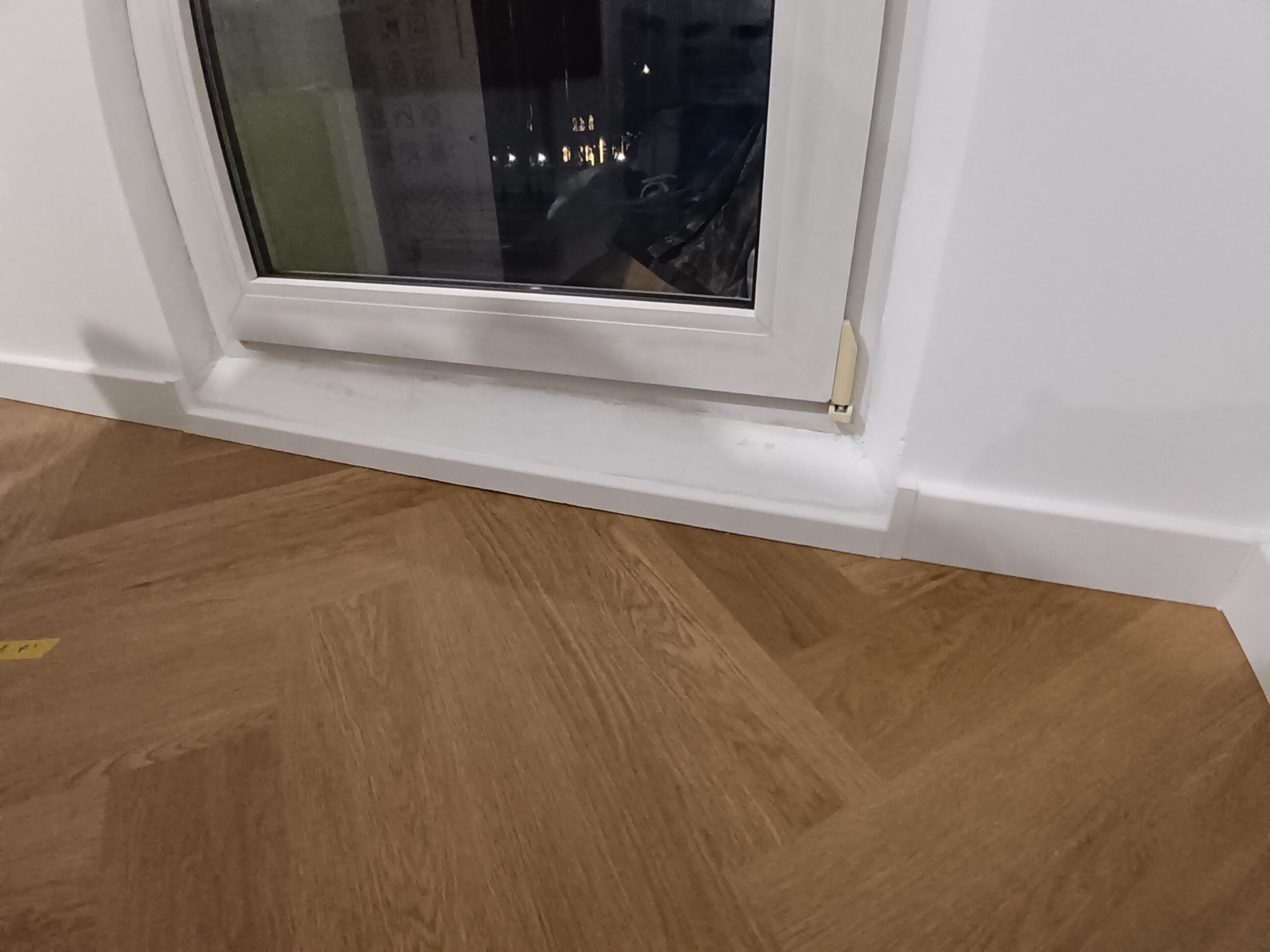 Parkiet ułożony w jodełkę francuską, widoczny fragment okna z białymi ramami i listwa przypodłogowa. Nowoczesne wykończenie podłogi w jasnym odcieniu drewna.