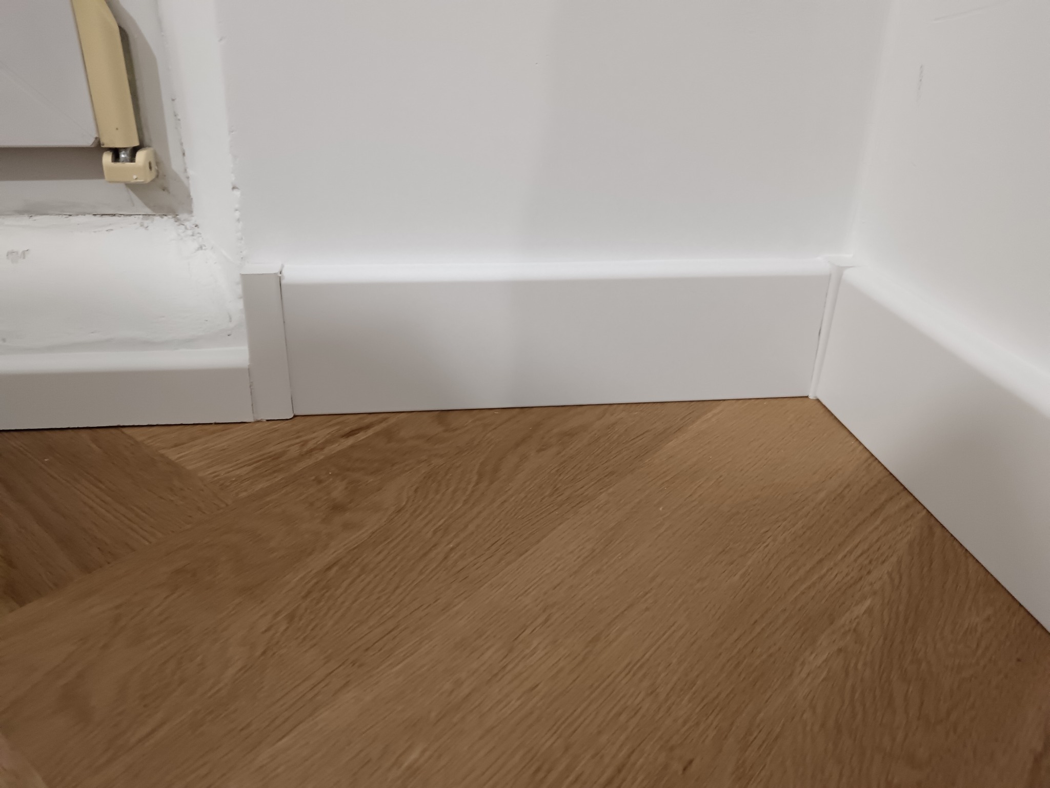 Jasne panele podłogowe ułożone w jodełkę, białe listwy przypodłogowe przy ścianie. Fragment grzejnika widoczny w lewym górnym rogu.