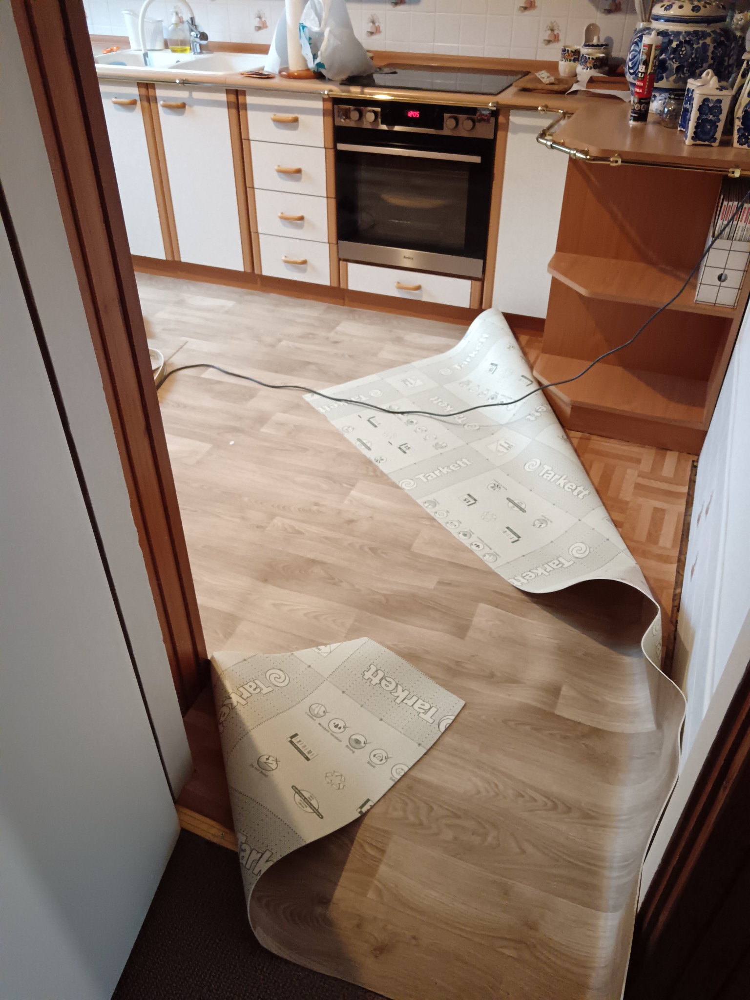 Fragment kuchni z rozłożonym panelem podłogowym Tarkett, przygotowanym do montażu. Widoczne szafki kuchenne w tle i fragment drzwi po lewej stronie.