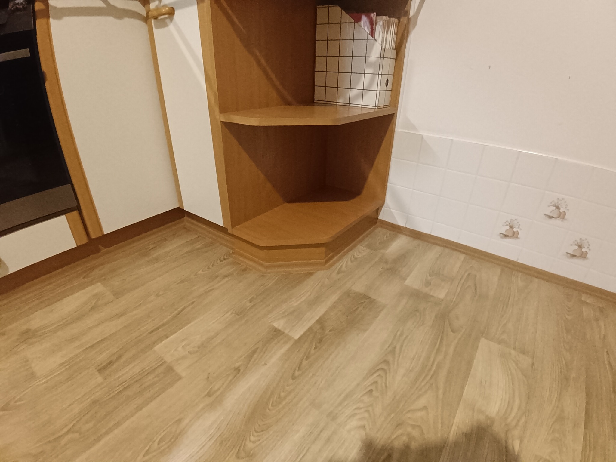 Jasne panele podłogowe ułożone w rogu pomieszczenia w Kosakowie, widoczne płytki na ścianie i drewniany regał narożny z półkami. Ciepłe oświetlenie.