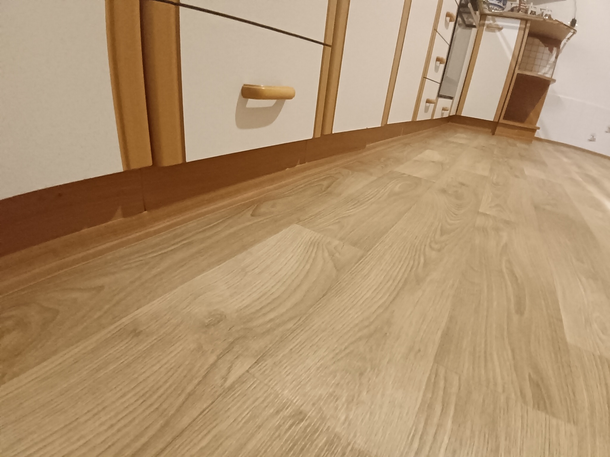 Jasne panele podłogowe z widoczną strukturą drewna, ułożone w pokoju z białymi meblami. Perspektywa z niskiego kąta podkreśla wzór paneli.