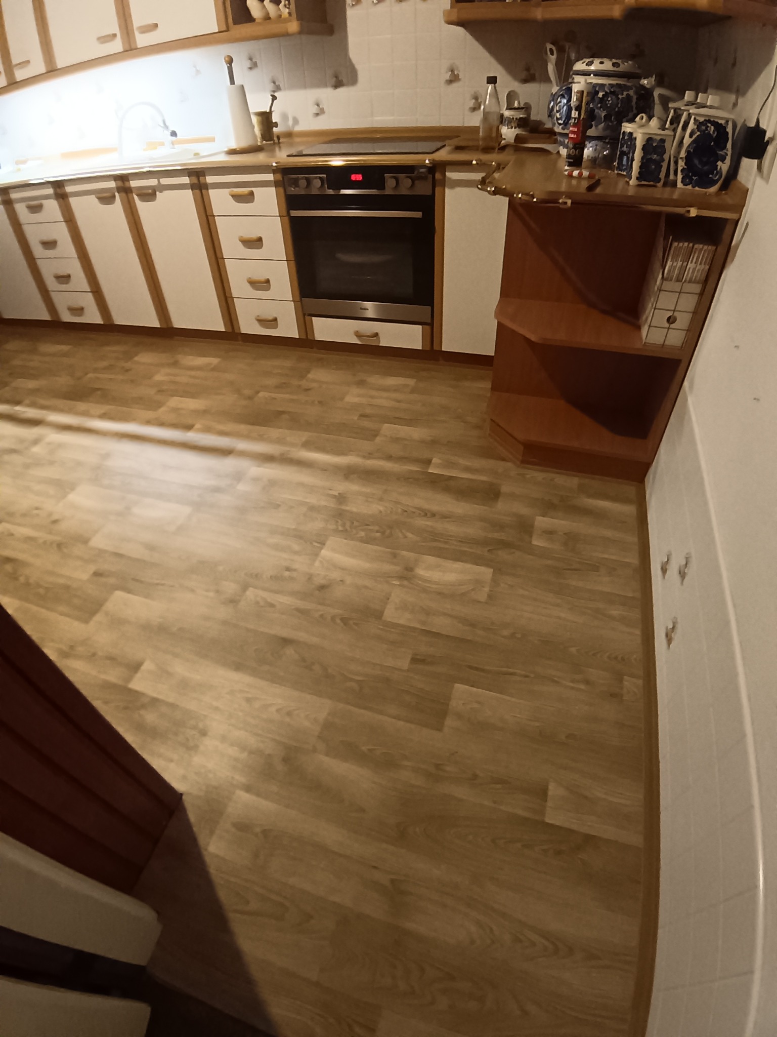 Jasna kuchnia z drewnianymi akcentami i nową podłogą z paneli. Widok na szafki, piekarnik i dekoracyjne naczynia. Ciepłe oświetlenie.