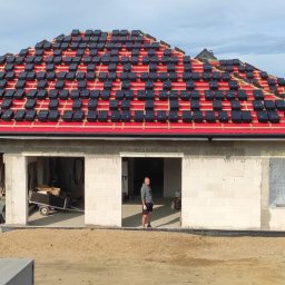 Usługi Więźba Dachowa SÖREN BRAND - Montaż czarnej dachówki na czerwonej membranie dachu garażu z białego bloczka. W tle zabudowa jednorodzinna i błękitne niebo.