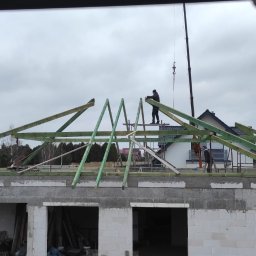 Usługi Więźba Dachowa SÖREN BRAND - Montaż drewnianej konstrukcji dachu na surowym budynku. Widoczni pracownicy i dźwig. Elementy więźby dachowej w kolorze zielonym. W tle zabudowa jednorodzinna.
