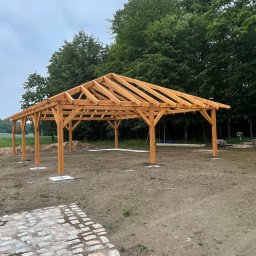 Usługi Więźba Dachowa SÖREN BRAND - Surowa, drewniana konstrukcja altany z widoczną więźbą dachową na betonowych podstawach, na tle zieleni i pochmurnego nieba. Widoczny fragment kamiennej ścieżki.