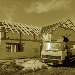 Usługi Więźba Dachowa SÖREN BRAND - Budowa domu z pustaków z drewnianą konstrukcją dachu w Nowogrodźcu. Na pierwszym planie samochód ciężarowy z dźwigiem. Dom w stanie surowym otwartym.