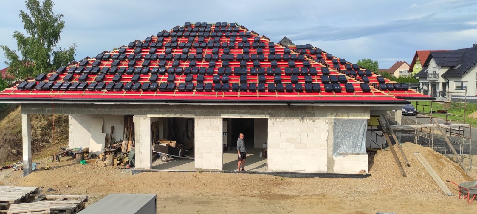 Montaż czarnej dachówki na czerwonej membranie dachu garażu z białego bloczka. W tle zabudowa jednorodzinna i błękitne niebo.