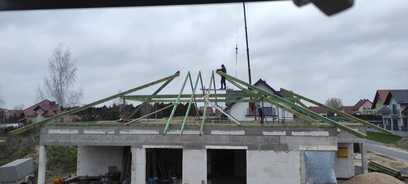 Montaż drewnianej konstrukcji dachu na surowym budynku. Widoczni pracownicy i dźwig. Elementy więźby dachowej w kolorze zielonym. W tle zabudowa jednorodzinna.