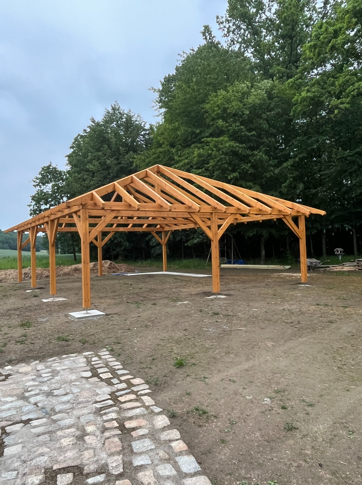 Surowa, drewniana konstrukcja altany z widoczną więźbą dachową na betonowych podstawach, na tle zieleni i pochmurnego nieba. Widoczny fragment kamiennej ścieżki.