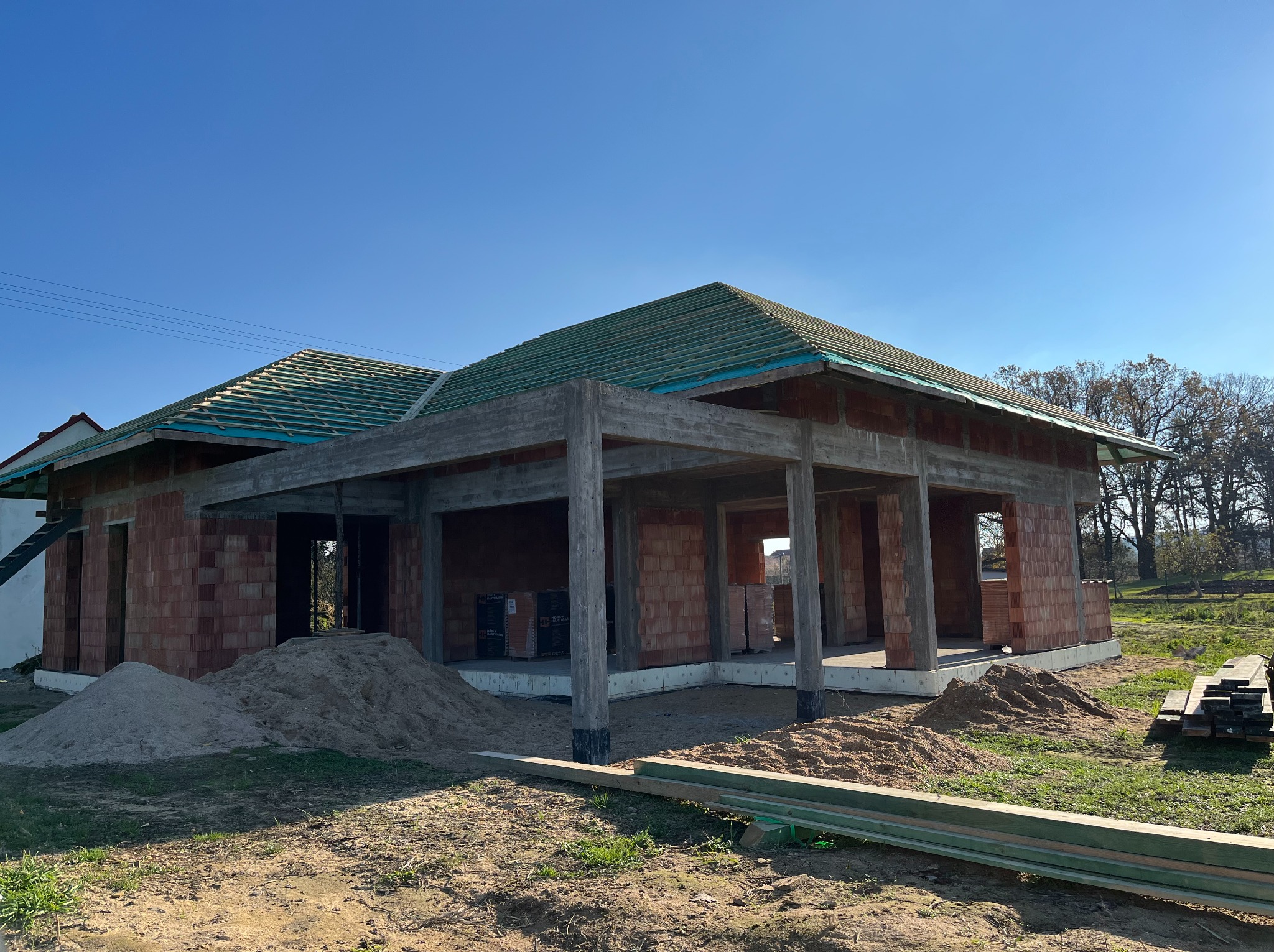 Budowa domu jednorodzinnego w stanie surowym otwartym z widoczną konstrukcją dachu i ścianami z cegły. Na pierwszym planie hałda piasku i deski.