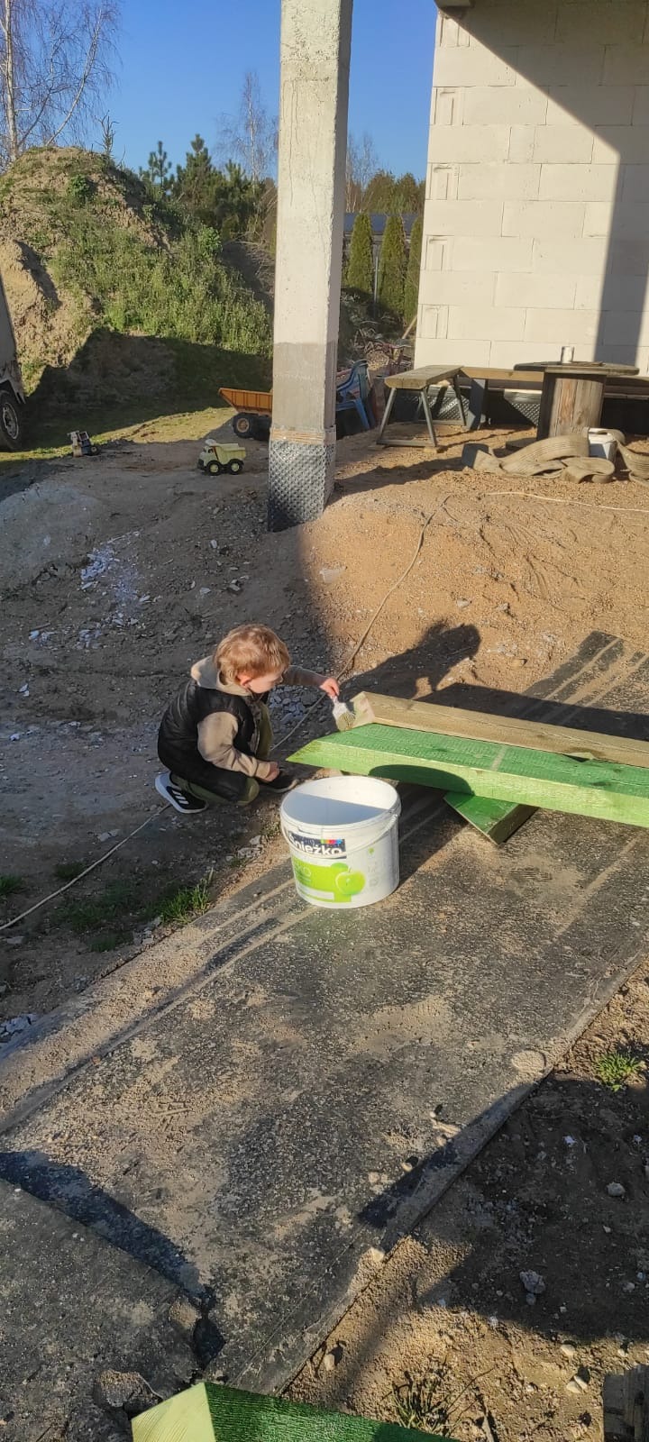 Chłopiec maluje drewno na zielono pędzlem przy wiadrze farby na placu budowy. W tle zabawki i fundamenty budynku.