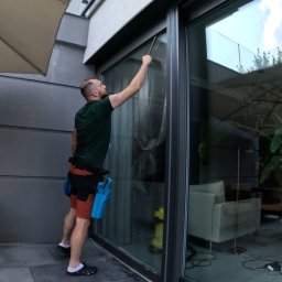 Piotr Kowalewski - Mężczyzna czyści okno panoramiczne w domu, używając ściągaczki. Widoczne narzędzia pracy i odbicie wnętrza domu w szybie. Praca wykonywana z zewnątrz.