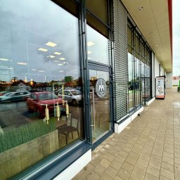 Piotr Kowalewski - Duże okna piekarni z widocznym odbiciem ulicy i wnętrza. Czyste szyby zapewniają dobrą widoczność ekspozycji i otoczenia. Widoczny chodnik i fragment elewacji.