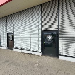 Piotr Kowalewski - Elewacja budynku z żaluzjami zewnętrznymi i dwoma wejściami z logo firmy. Widok z poziomu chodnika, neutralne oświetlenie dzienne.
