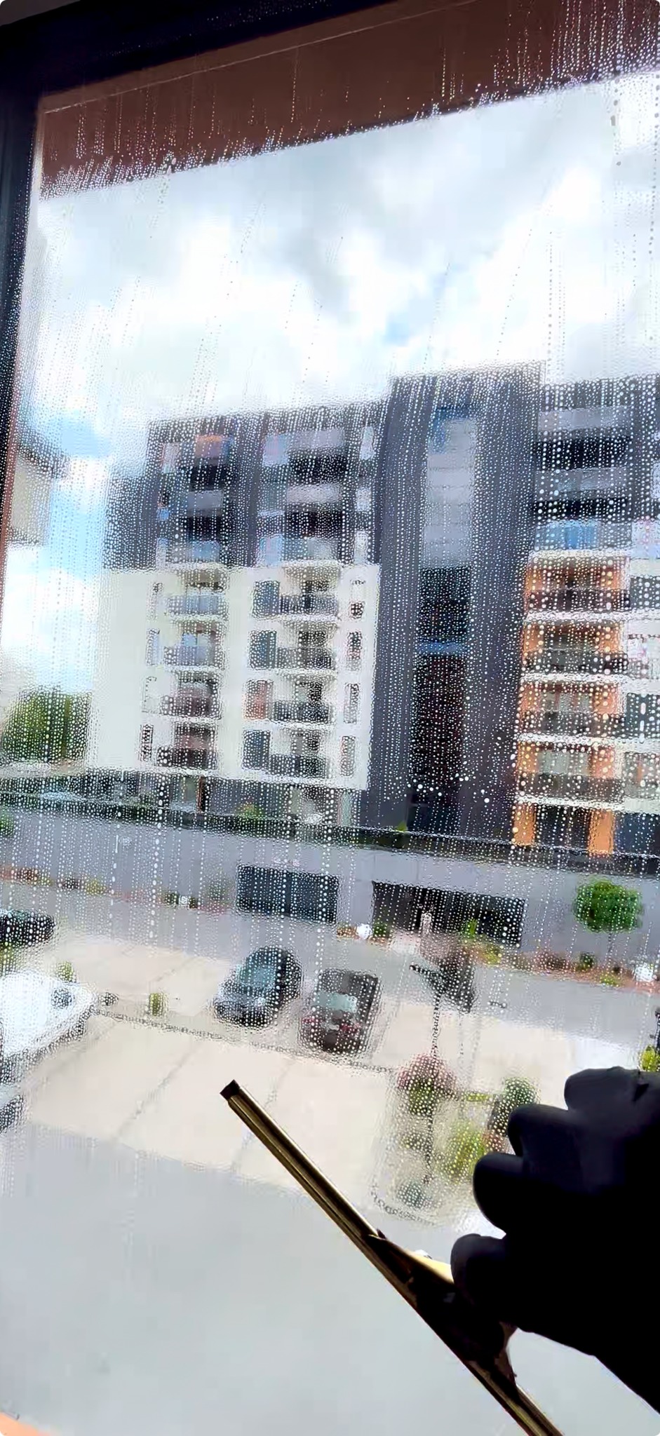 Okno w trakcie czyszczenia z widokiem na budynki mieszkalne i parking. Używany ściągacz do szyb w dłoni w czarnej rękawicy.