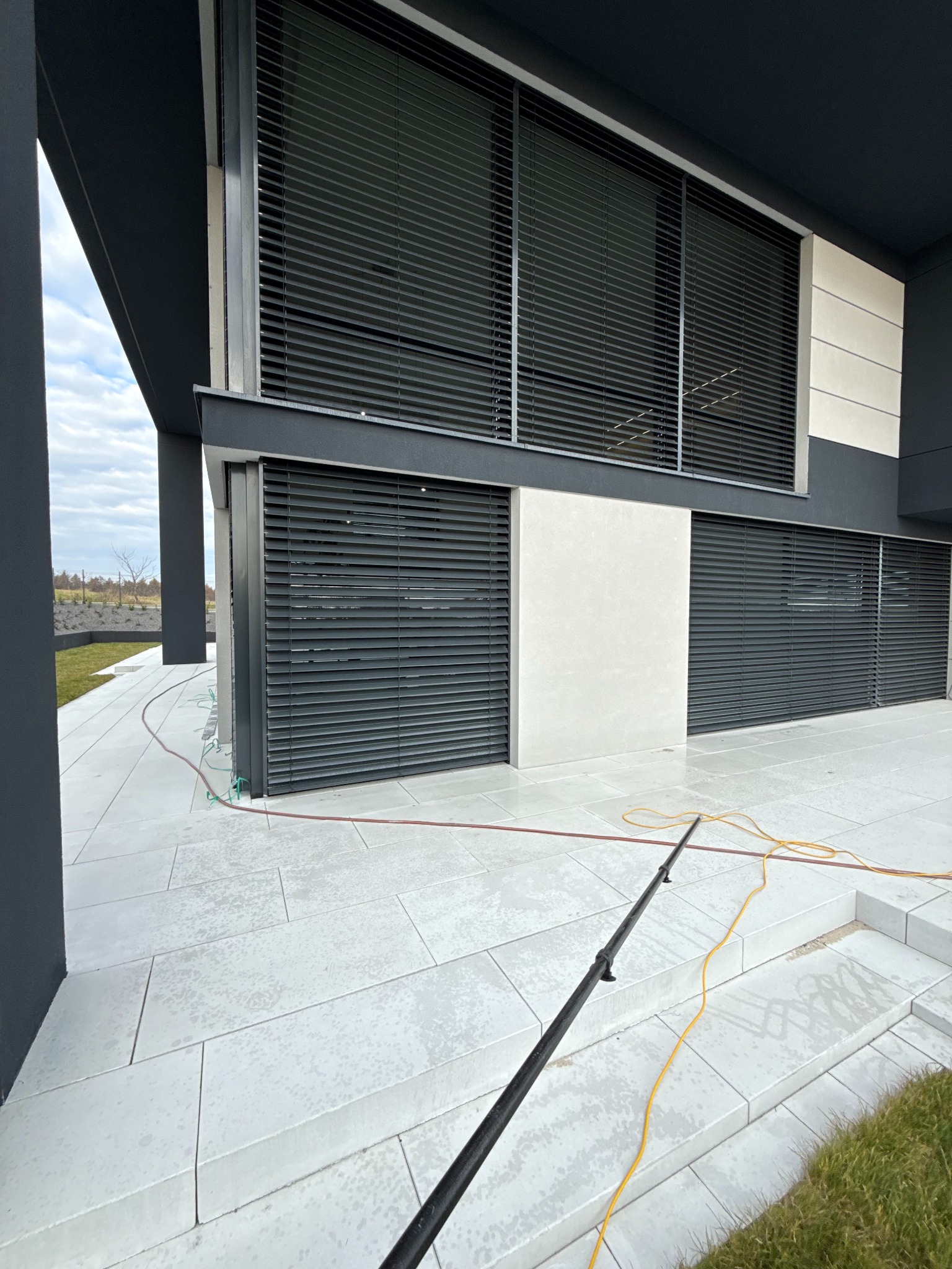 Nowoczesny budynek z szarymi żaluzjami zewnętrznymi, czyszczony lancą teleskopową. Ślady wody na jasnej posadzce. Widok z dołu, lekki kąt.