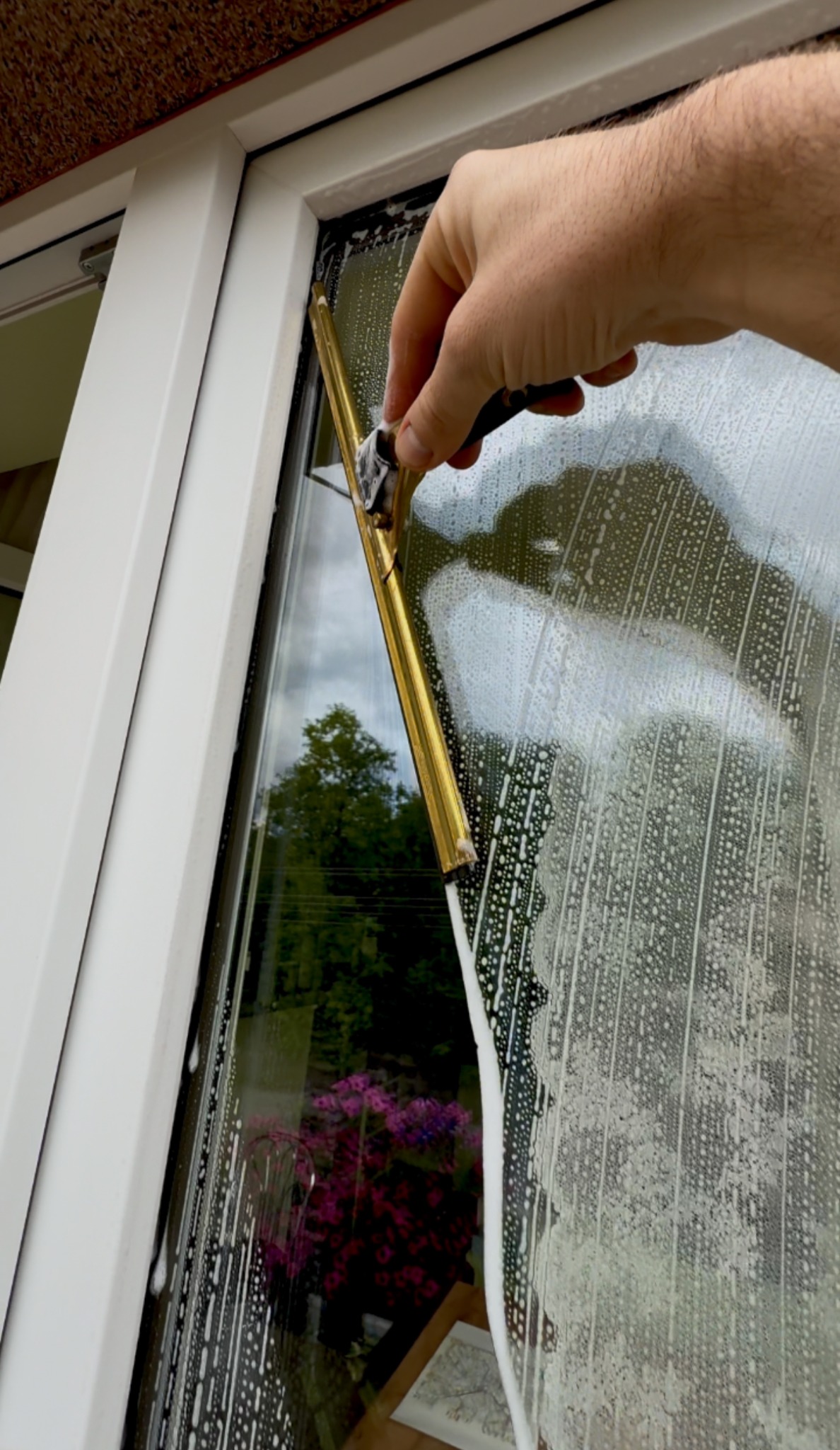 Mycie okna z użyciem ściągaczki z gumą, widoczne smugi piany. W tle zieleń drzew i kwiaty odbijające się w szybie. Biała rama okienna.