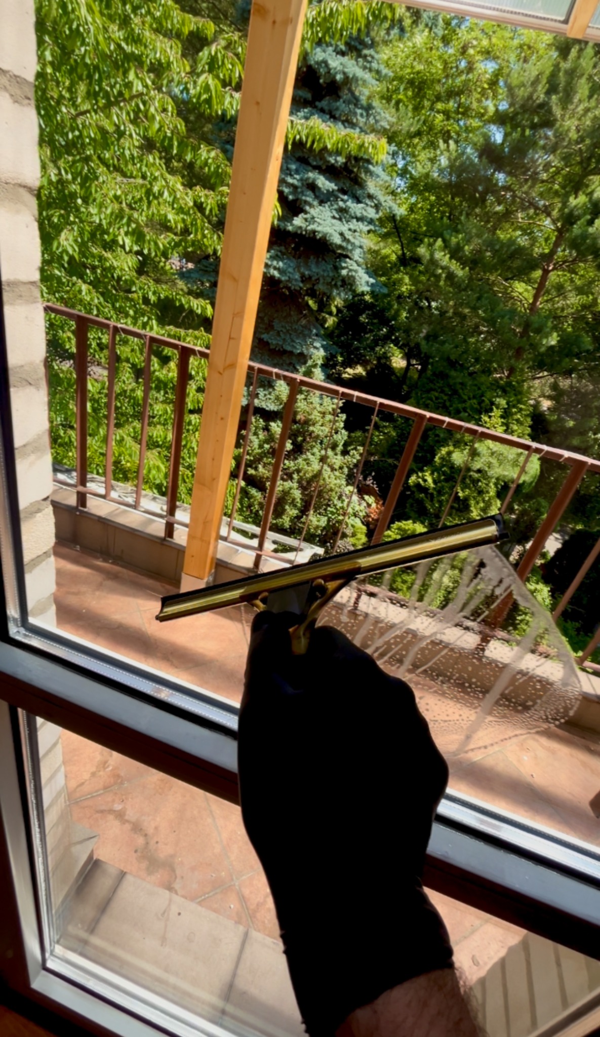 Czyszczenie okna z zewnątrz za pomocą ściągaczki, widok z balkonu z zielonym tłem. Dłoń w czarnej rękawicy prowadzi narzędzie po szybie.