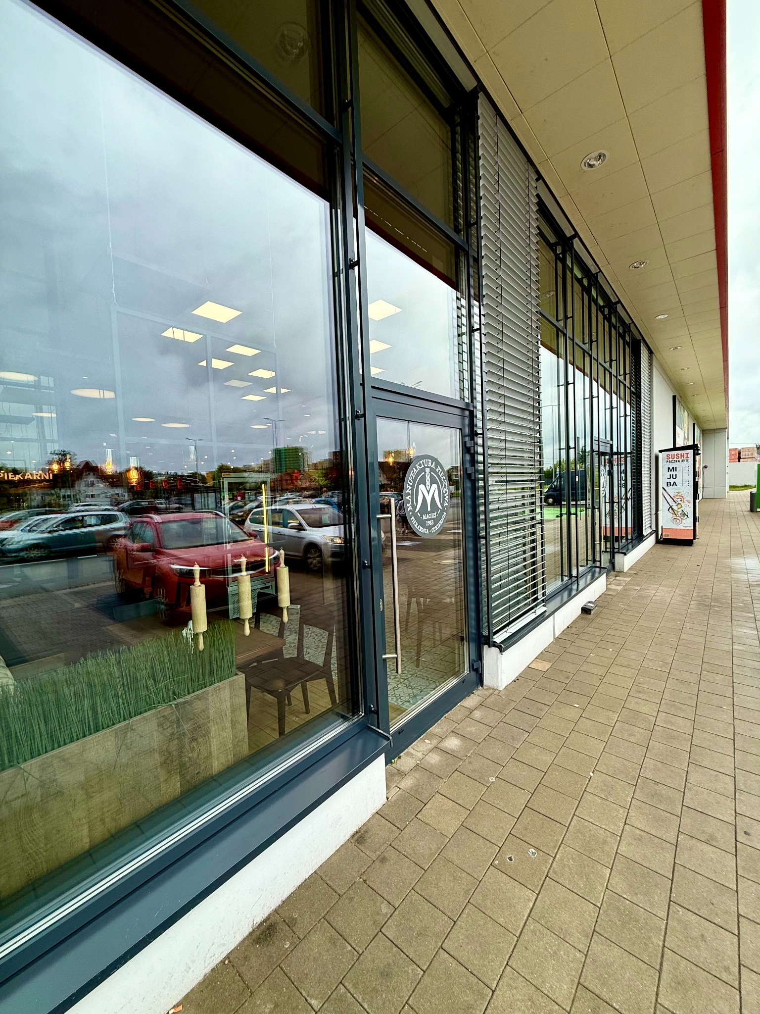 Duże okna piekarni z widocznym odbiciem ulicy i wnętrza. Czyste szyby zapewniają dobrą widoczność ekspozycji i otoczenia. Widoczny chodnik i fragment elewacji.