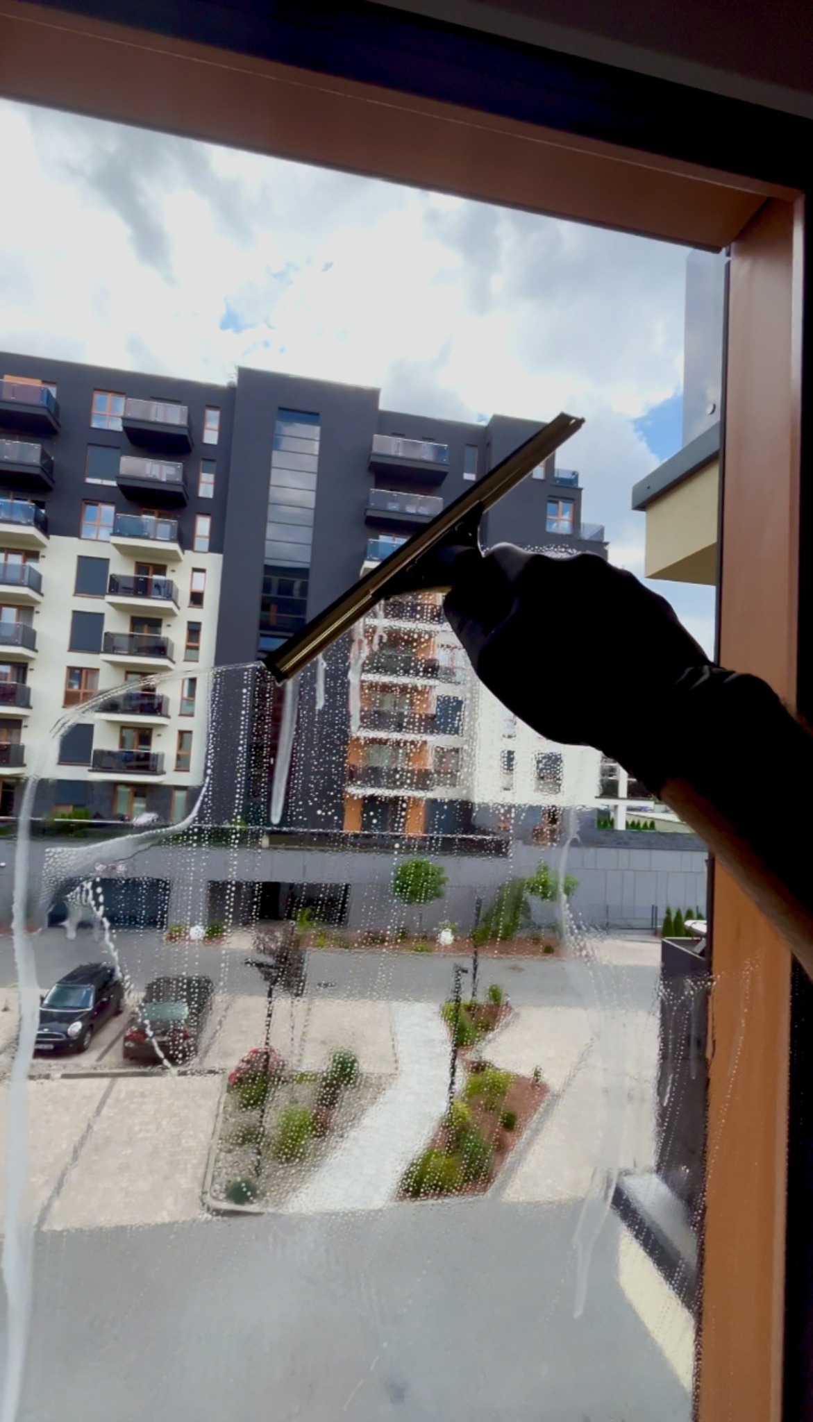 Czyszczenie okna z zewnątrz budynku mieszkalnego za pomocą ściągaczki. Widoczne smugi piany na szybie i odbicie otoczenia.