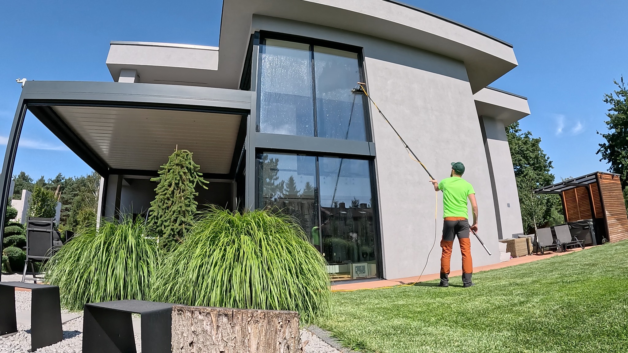 Pracownik w zielonej koszulce czyści okna nowoczesnego domu za pomocą długiej lancy. Widoczne ślady wody na szybach, zadbany trawnik i elementy architektury ogrodowej.