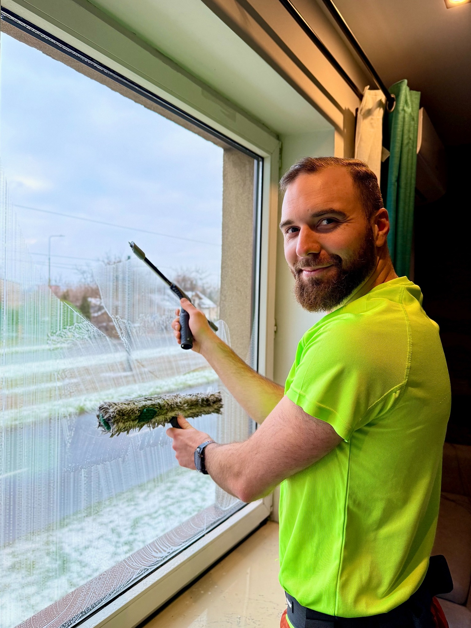 Uśmiechnięty mężczyzna w neonowej koszulce myje okno za pomocą ściągaczki i myjki. Widoczne smugi piany na szybie i fragment ulicy za oknem.