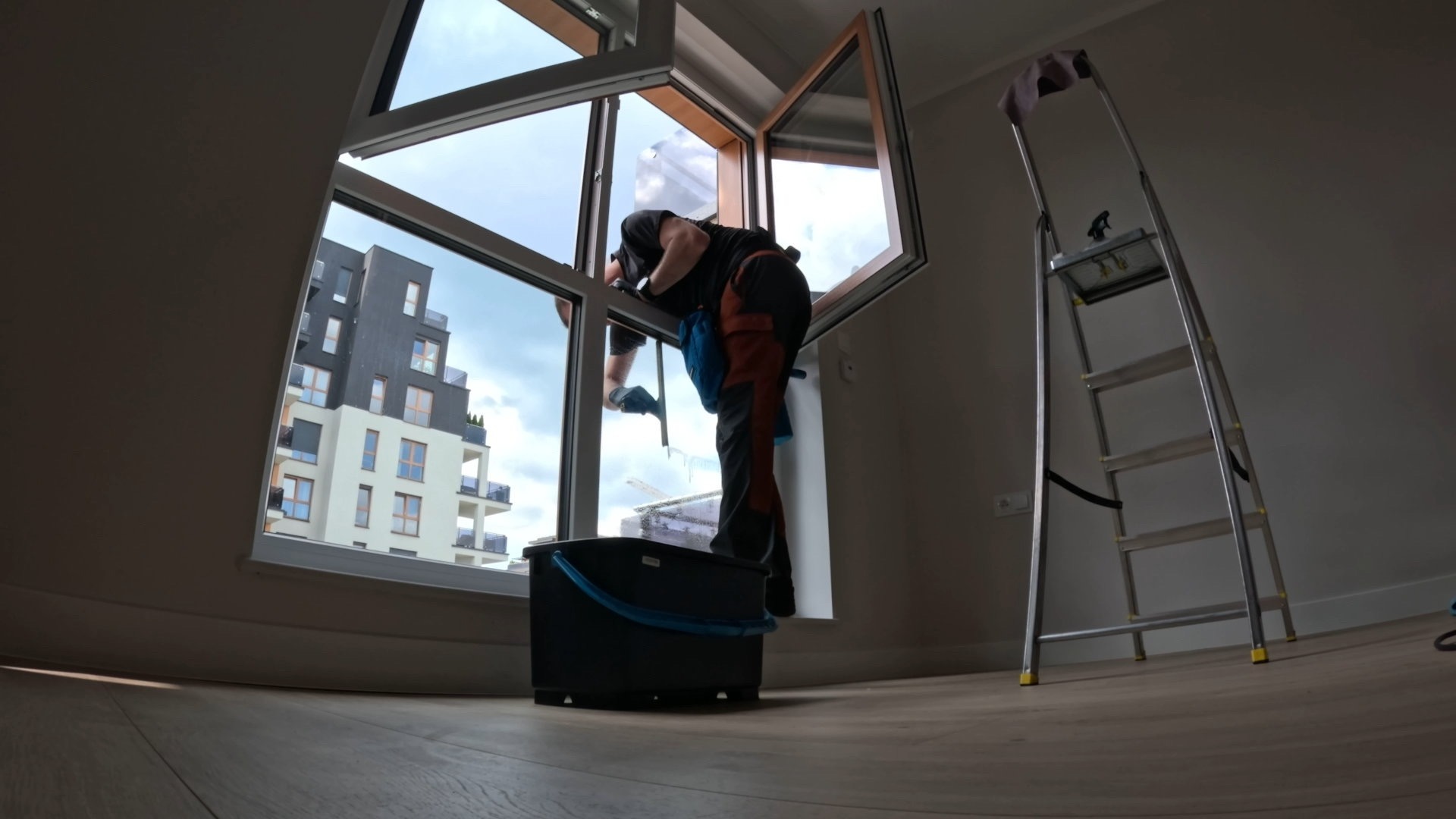 Pracownik w trakcie czyszczenia okien w mieszkaniu, widok z dołu. Na zewnątrz widoczny budynek mieszkalny. Obok drabina i wiadro z akcesoriami.