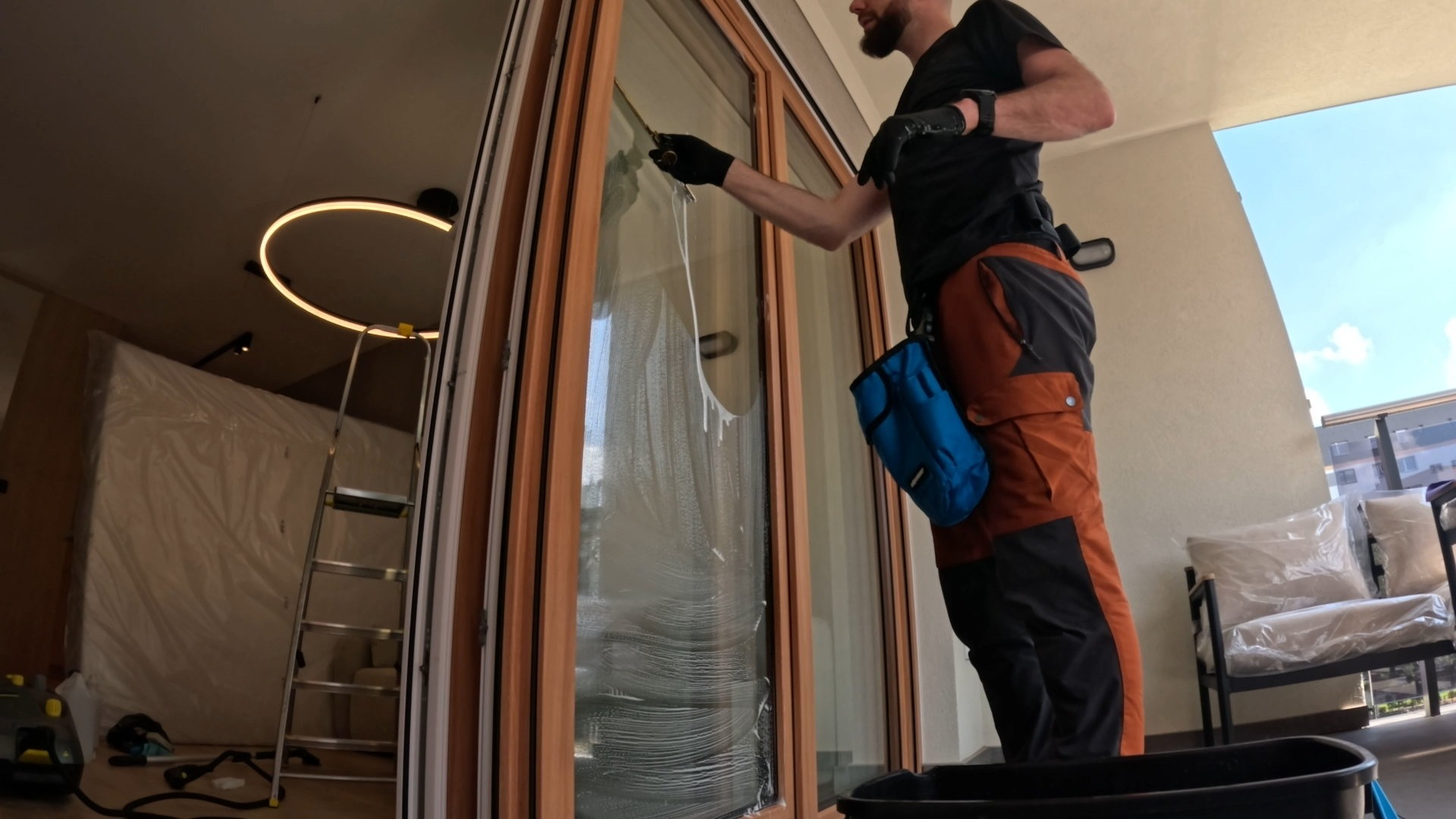 Mężczyzna w czarnej koszulce czyści okno od wewnątrz, używając ściągaczki. Widoczne smugi wody na szybie, drabina i meble zabezpieczone folią w tle.