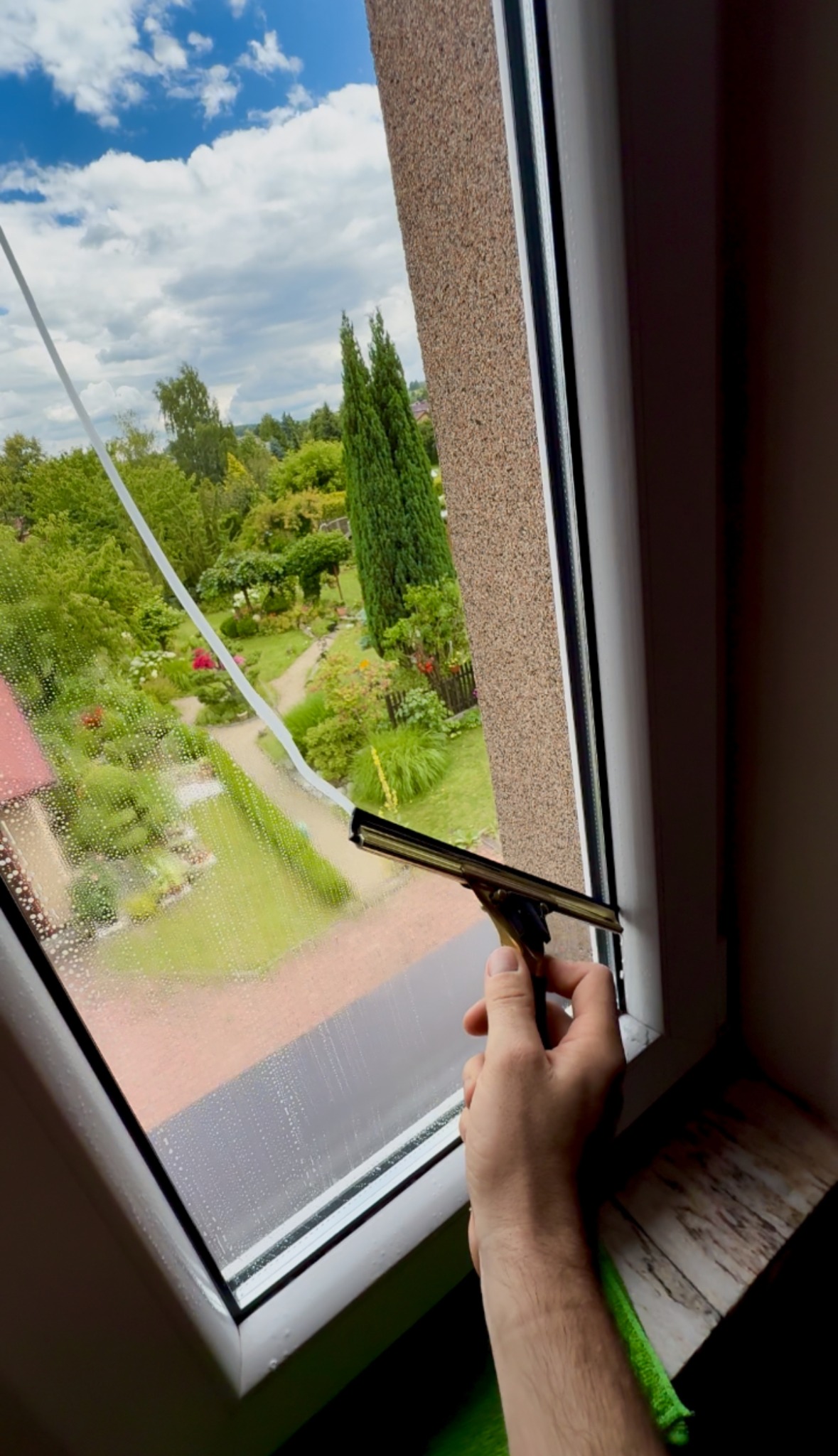 Czyszczenie okna w Będzinie za pomocą ściągaczki. Widok na zielony ogród zza szyby. Drobne krople wody na szkle. Biała rama okienna.