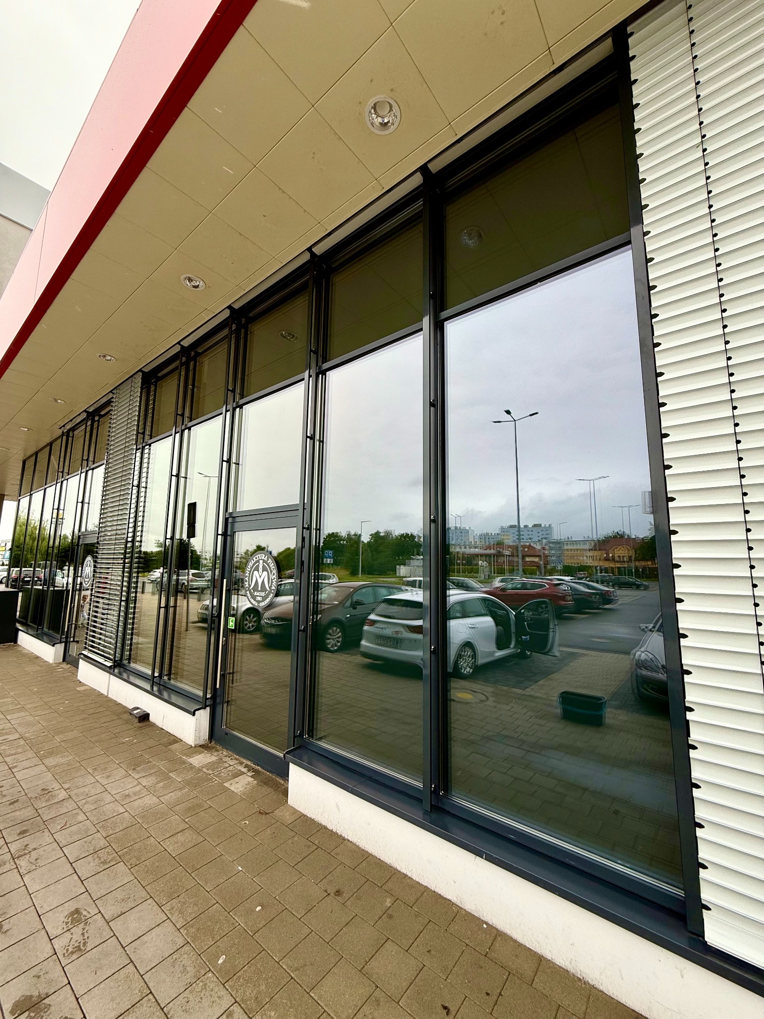 Błyszczące, umyte okna witryny sklepowej z czarnymi ramami, odbijające parking i budynki. Widoczne rolety zewnętrzne i chodnik z kostki brukowej.