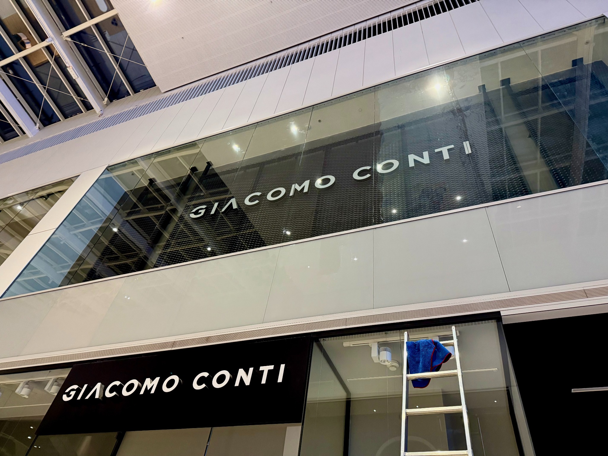 Mycie witryny sklepowej Giacomo Conti w galerii handlowej. Na drabinie leży niebieska ścierka. Widoczne logo sklepu na szybie i nad wejściem.