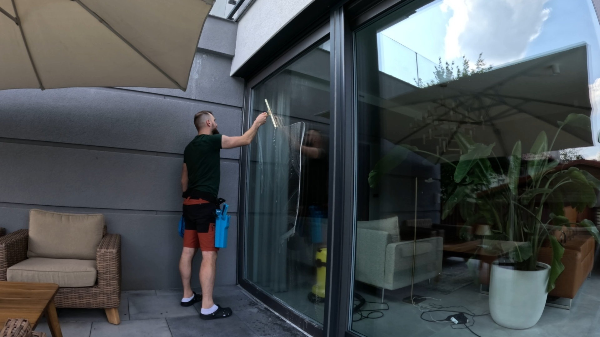 Mężczyzna w trakcie mycia okna na tarasie nowoczesnego domu. Widoczne narzędzia i odbicie wnętrza domu w czystej szybie.