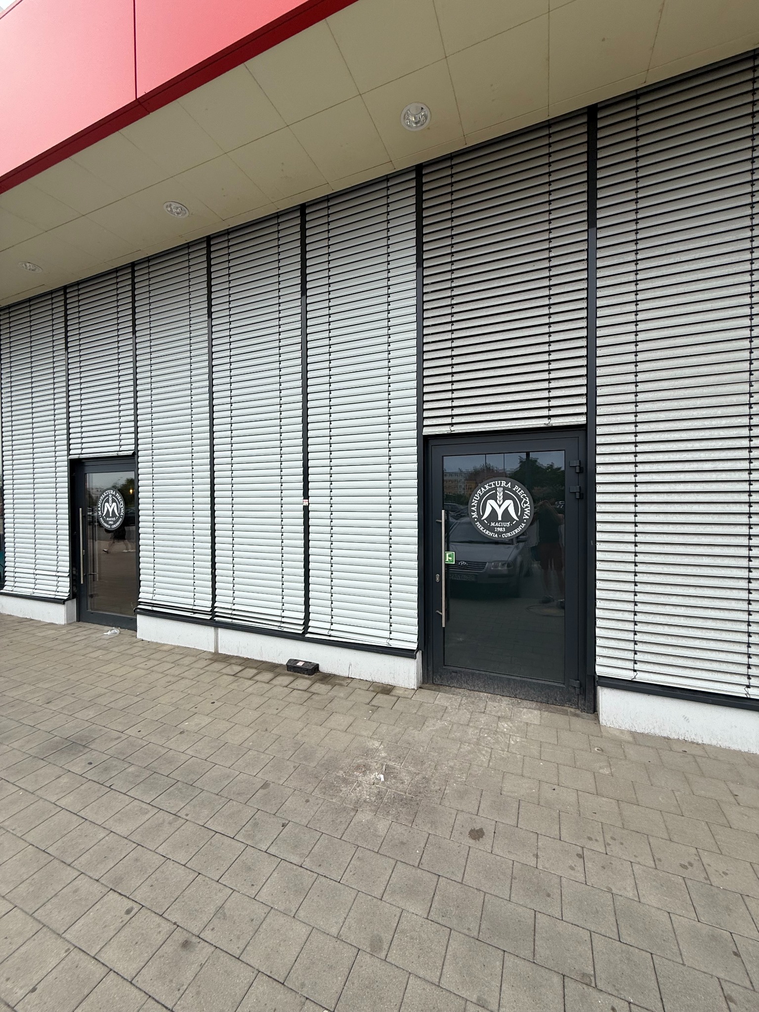 Elewacja budynku z żaluzjami zewnętrznymi i dwoma wejściami z logo firmy. Widok z poziomu chodnika, neutralne oświetlenie dzienne.